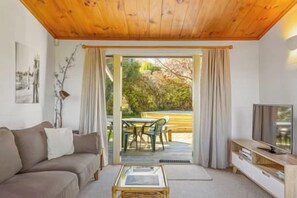 A living room with a sofa, coffee table, and a television. There is a sliding glass door leading to an outdoor area with a table and chairs. - Bellbird Cottage - private large section, with lake and mountain views. Perfect escape to relax (Taupo)