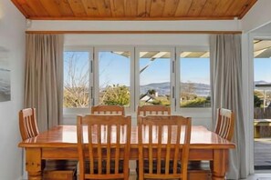A dining area with a wooden table and chairs, large windows with curtains, and a view of the outdoors. - Bellbird Cottage - private large section, with lake and mountain views. Perfect escape to relax (Taupo)