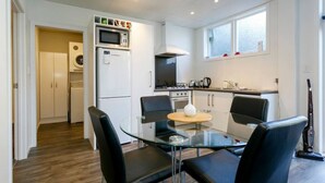 Cocina compacta con microondas, estufa y lavabo, una mesa de vidrio con cuatro sillas, y un ventanal que proporciona luz natural.