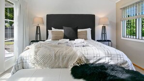 Een slaapkamer met een groot bed, twee nachtkastjes met lampen, een raam met zonwering en uitzicht op groen.