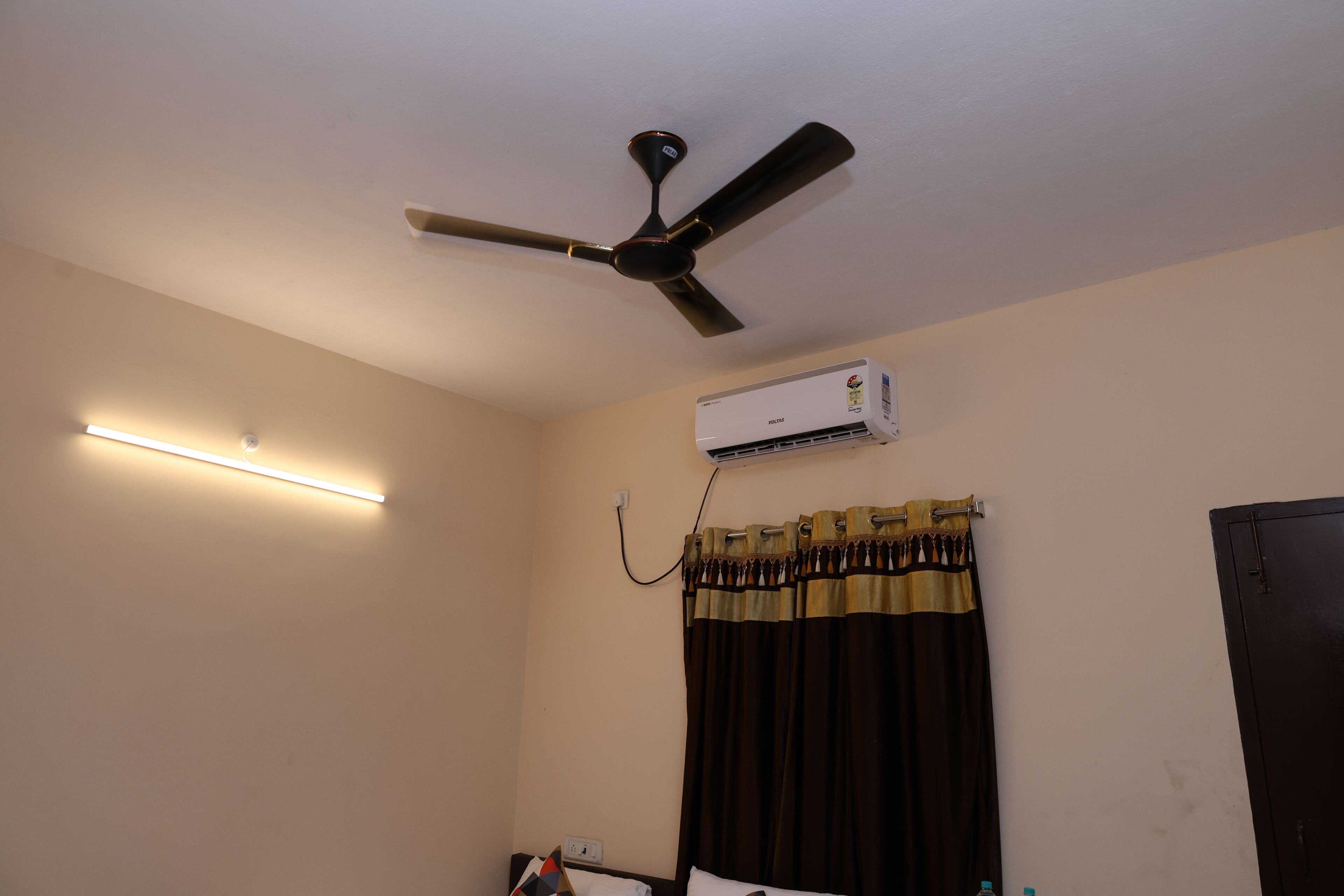 Habitación de hotel con ventilador de techo, aire acondicionado, mesita de noche con lámpara y ventana con cortinas.