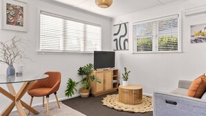 A modern living room with a glass dining table, a sofa, a TV on a wooden cabinet, and potted plants.