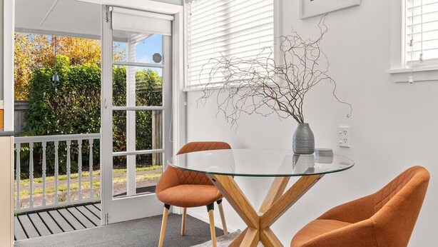 A modern dining area with a glass table, two orange chairs, a vase with branches, and a view through a sliding glass door to a garden.