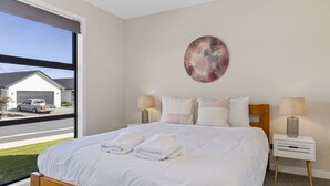 Une chambre avec un grand lit, deux tables de chevet avec des lampes, une œuvre d’art murale, et une vue sur une maison et une rue, visible à travers une porte coulissante.