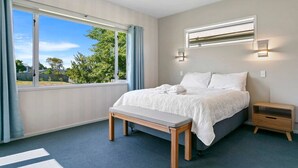 Een slaapkamer met een groot raam, een bed met witte lakens, een nachtkastje en een bankje.