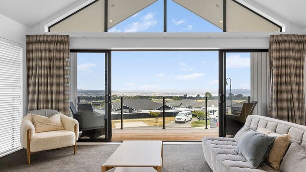 A modern living room with a sofa, armchair, and coffee table, featuring a large sliding glass door leading to a balcony with a view of a residential area. - Blue Dun Retreat | Relaxing, Modern Getaway Near Lake & Town (Taupō)