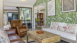Ein modernes Wohnzimmer mit Ledersessel, weißes Sofa mit gemusterten Kissen, Couchtisch aus Holz und einer Wand mit grünem Blattmuster.
