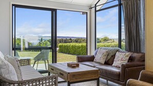 Ein modernes Wohnzimmer mit Ledersofa, Couchtisch aus Holz und Korbstuhl. Große Fenster bieten Blick auf einen üppigen Garten und ferne Hügel.