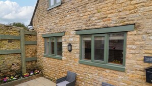 A stone cottage with two windows, wicker furniture, and a flower bed. - Triumph Cottage (Chipping Norton)