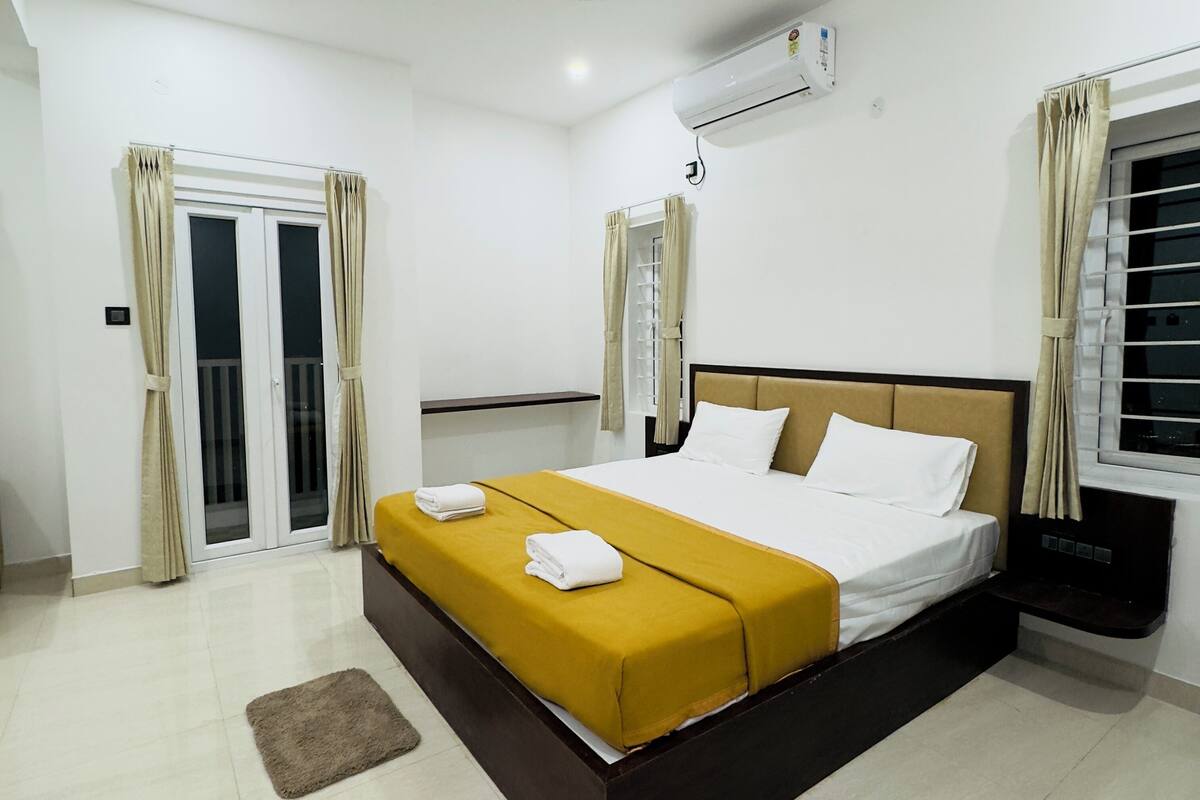 Quarto de hotel com cama, mesa de cabeceira, ar condicionado, janela com cortinas e porta de correr.