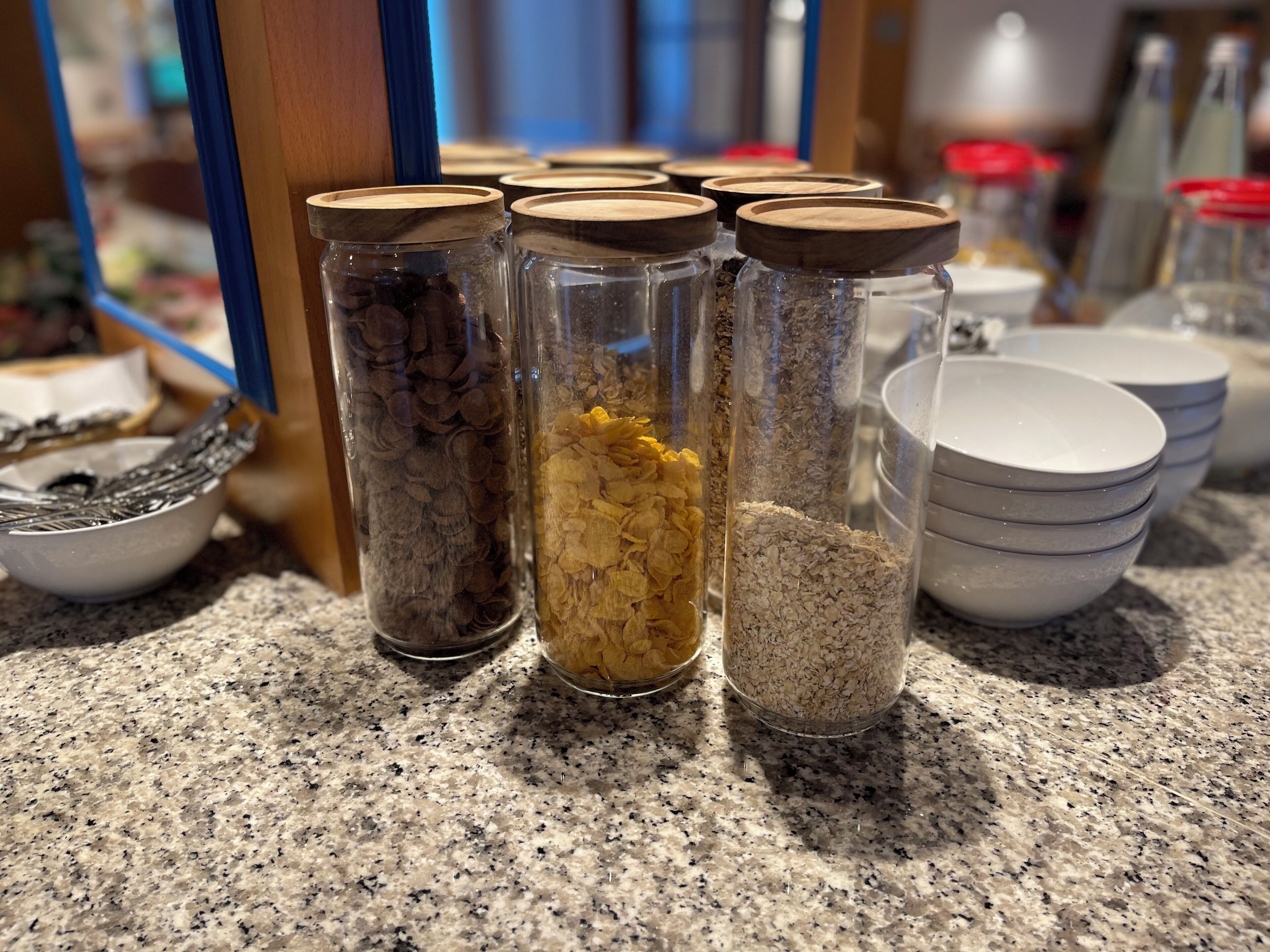 A kitchen counter with jars containing various grains and seeds, a bowl of utensils, and stacked bowls.