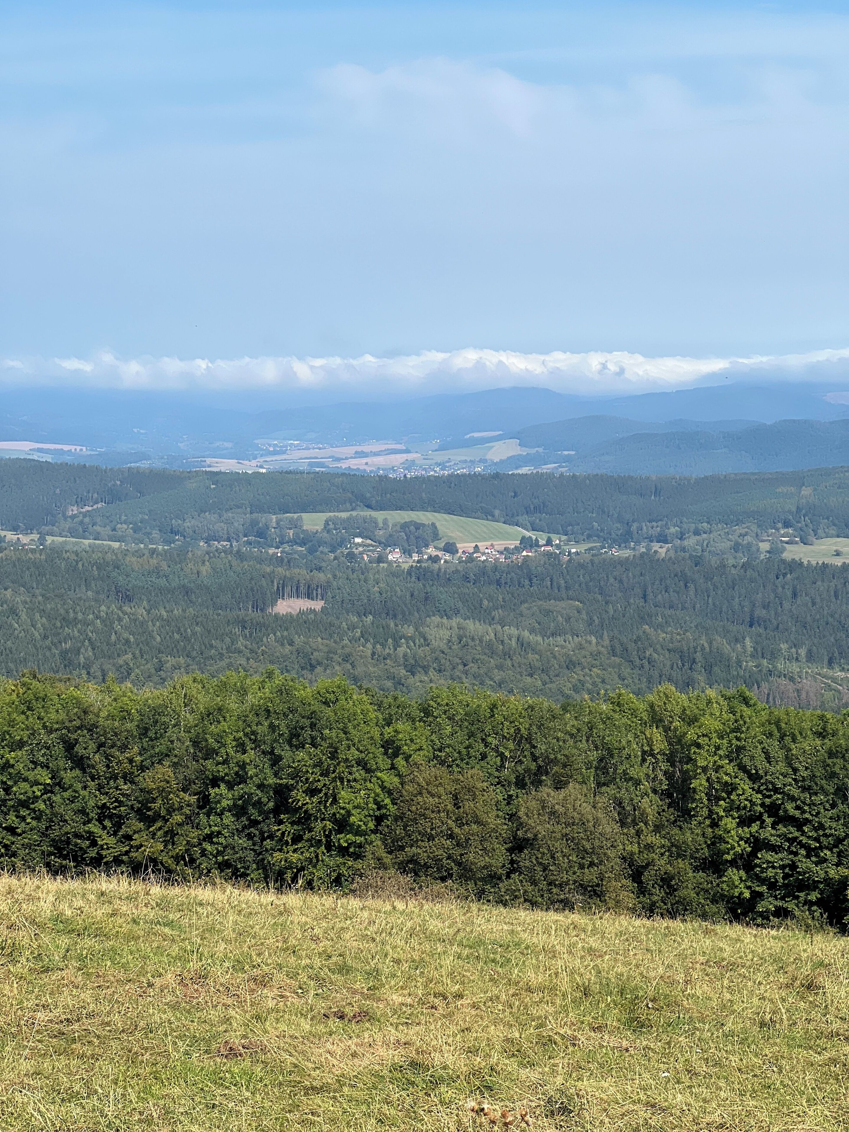 Vista panorâmica de uma paisagem montanhosa com um vale arborizado, um pequeno povoado e montanhas distantes sob um céu claro.