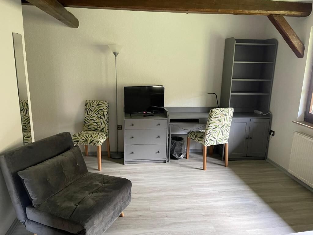 A compact living space with a sofa, two patterned chairs, a desk with a TV, and a shelving unit.