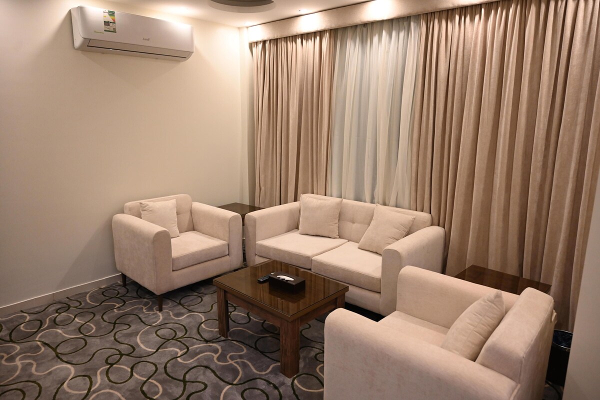 A hotel room with a beige sofa set, a wooden coffee table, and a large window with curtains.