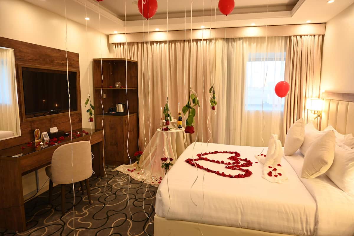 Quarto de hotel com cama, escrivaninha, cadeira, televisão, guarda-roupa e janela com cortinas.