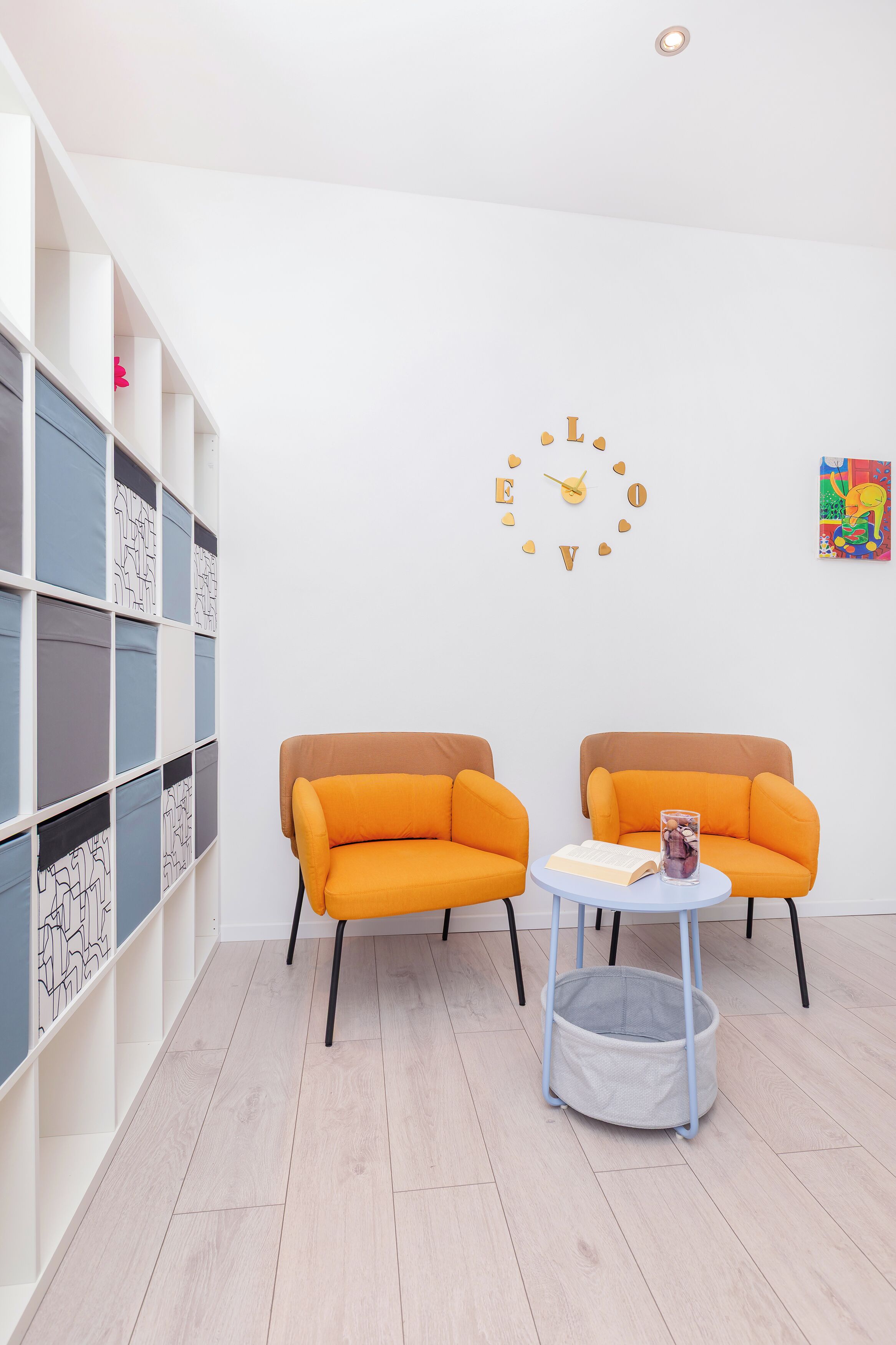Una habitación con dos sillones naranjas, una pequeña mesa redonda con un libro y un vaso, y una estantería con paneles azules y blancos.