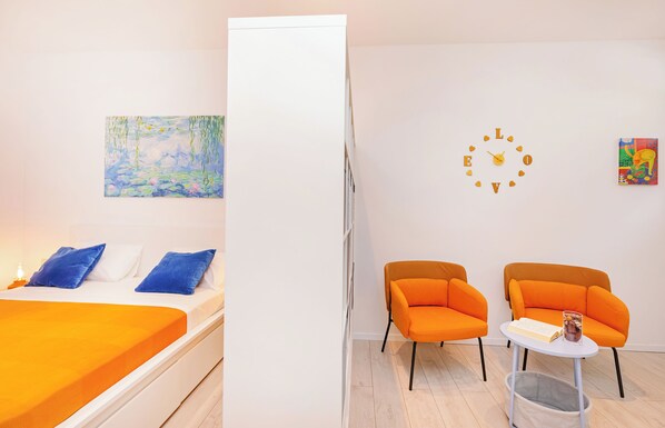 A room with a bed, two orange chairs, a small table with a book and a glass, and a wall clock. - Bis Studio Apartment 4 (Dubrovnik)