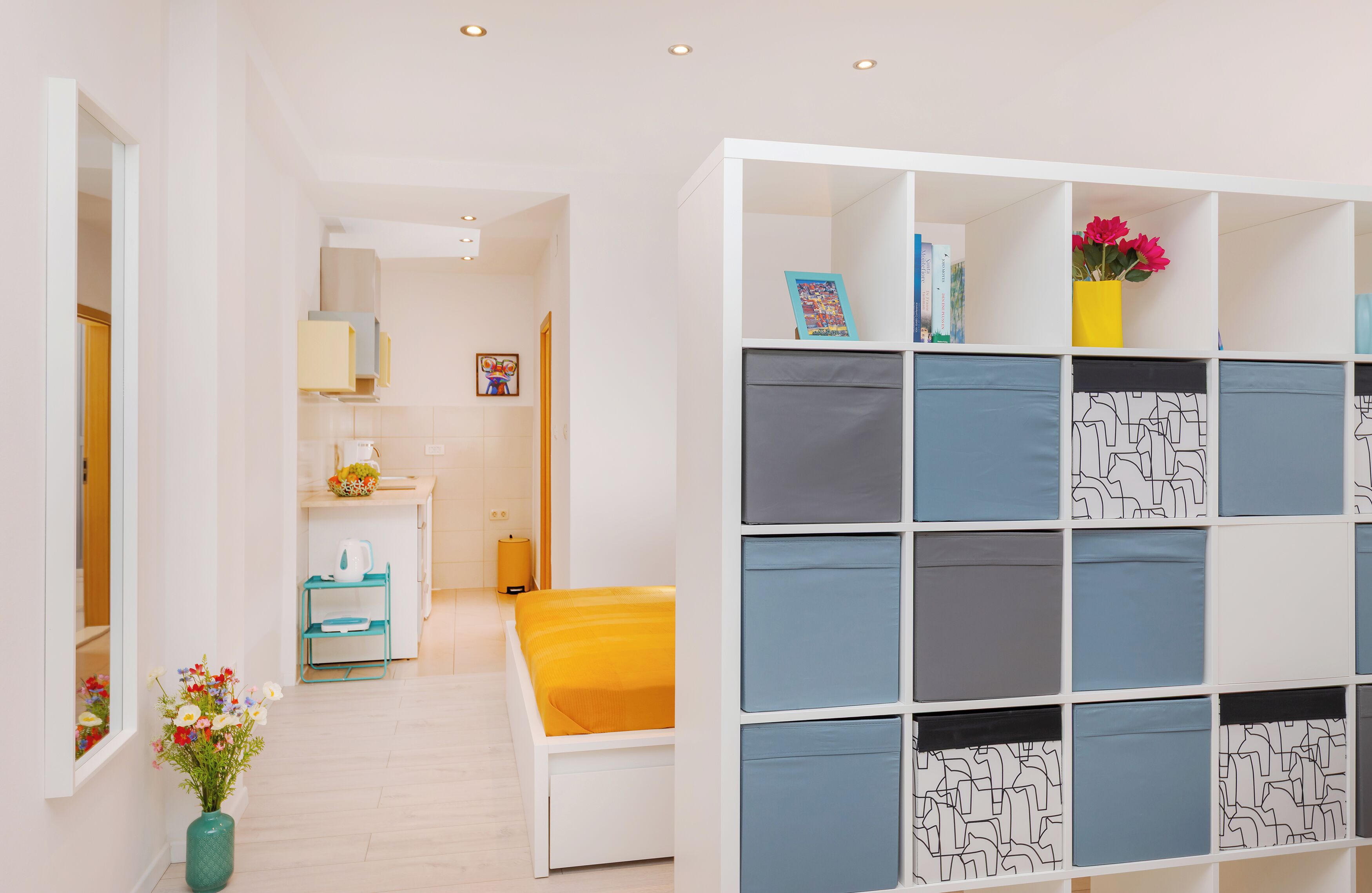 Habitación de hotel moderna con una cama blanca, un pequeño escritorio y una estantería con cajas de almacenamiento estampadas.