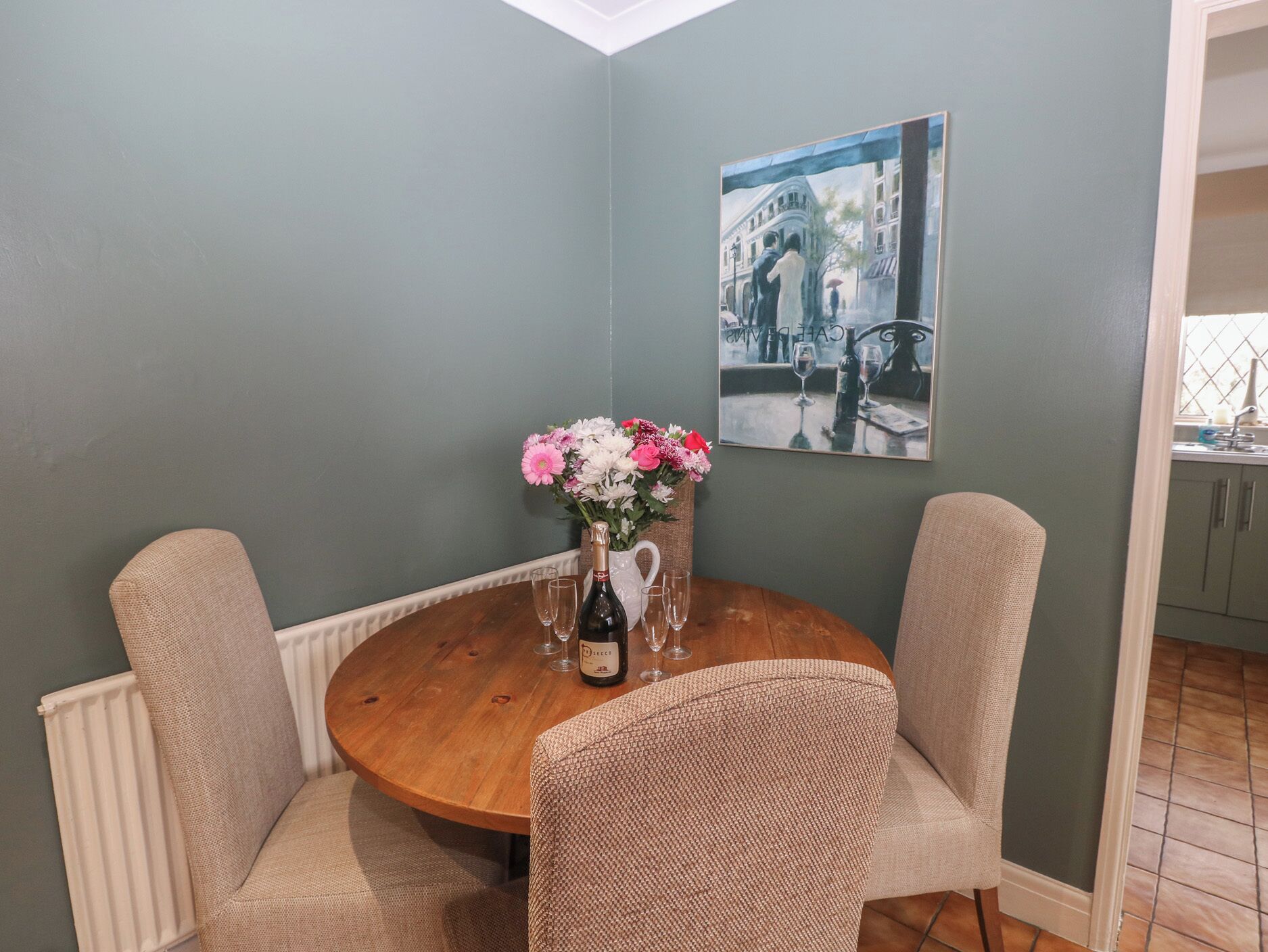 A dining area with a round wooden table, two beige chairs, a vase with flowers, and a bottle of wine.