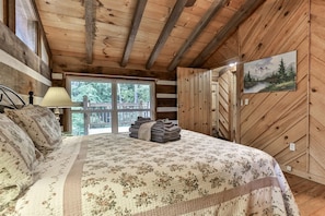 Quarto de cabana de madeira com cama, abajur, pintura e vista para o exterior.