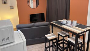 A modern living room with a sofa, a TV, a dining table with stools, and a small kitchen area with a microwave. - Fully-equipped studio just a stone's throw from the Château de Lunéville (Lunéville)