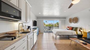 A modern kitchen with a built-in oven, a dining area with a table and chairs, a bedroom with a bed and a nightstand, and a balcony with a view of palm trees.