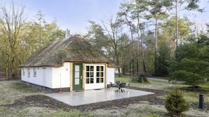 Een klein wit huis met rieten dak, een groene deur en een terras met zitplaatsen buiten.