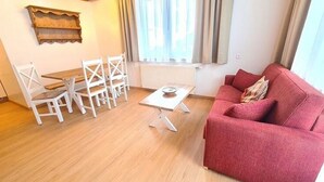 A living room with a wooden floor, a red sofa, white chairs, a small table, and a window with curtains. - Cosy apartment in Bad Kleinkirchheim (Bad Kleinkirchheim)