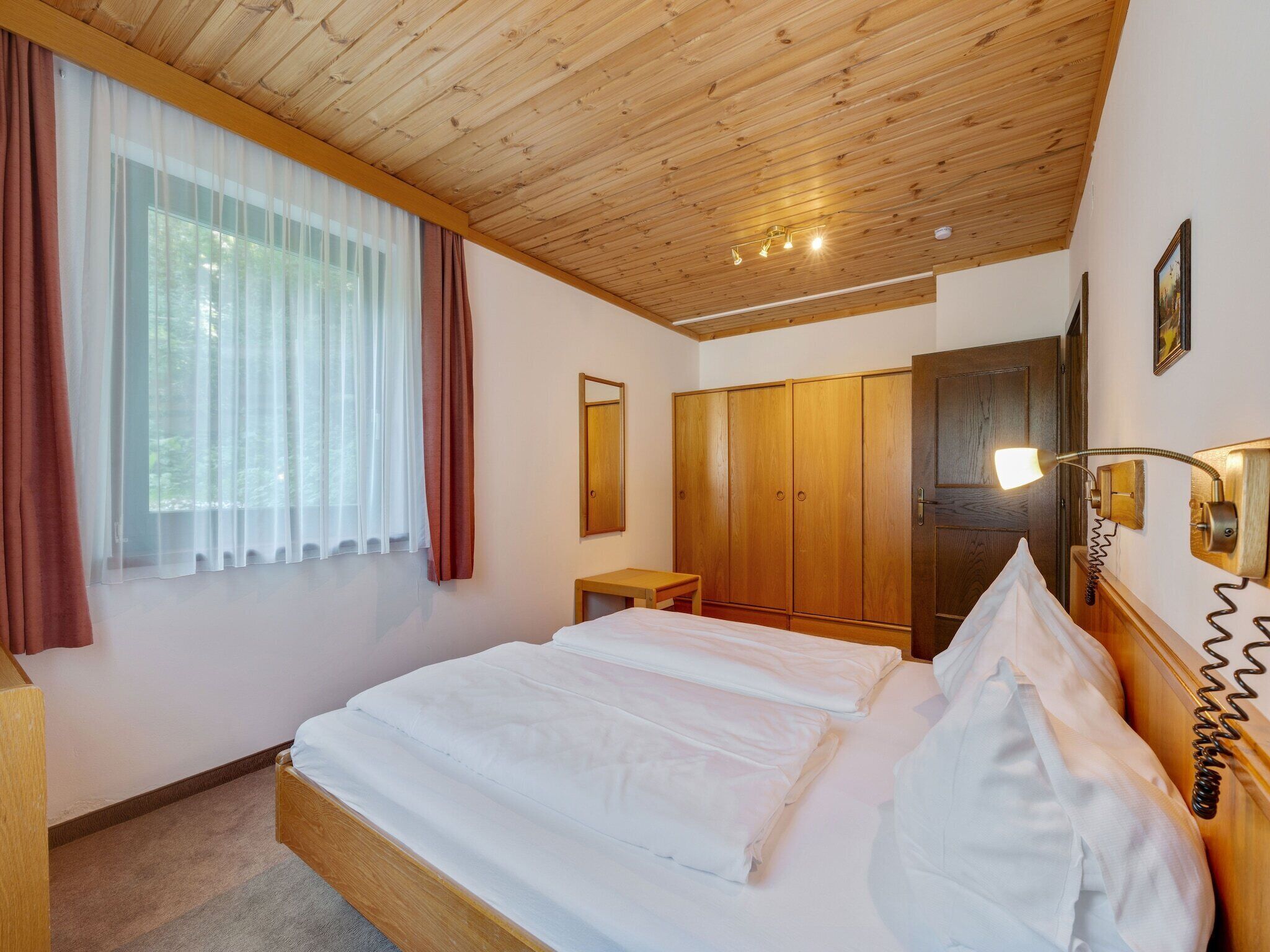 Un dormitorio con techo de madera, una cama con ropa blanca, un armario de madera, una mesita de noche y una ventana con cortinas traslúcidas.