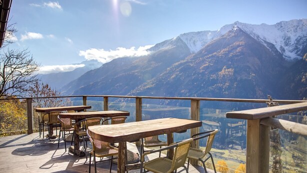 An outdoor terrace with wooden tables and chairs, overlooking a mountainous landscape.
