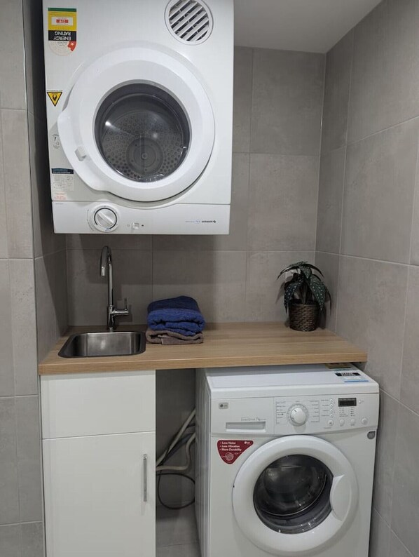 A compact laundry area with a top-loading washing machine and a front-loading dryer, a sink, and a small shelf with folded towels. - Supremely Spacious @Kingston (Kingston)