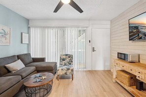 Een moderne woonkamer met een bank, een gestreept fauteuil, een bijzettafeltje met zeesterren, een plafondventilator, een televisie en een houten kast.