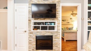 A living room with a stone fireplace, a mounted TV, and a bed in the background.
