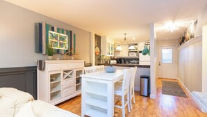 A compact kitchen with white cabinetry, a dining table, and chairs. There is a trash can and a rug near the door.