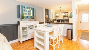 A compact kitchen with white cabinetry, a dining table, and chairs. There is a trash bin and a small potted plant on the counter.