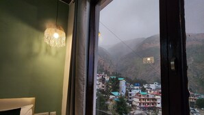 A room with a bed, a window with a view of a town and mountains, and a hanging light fixture. - Hotel Mahadev (Dharamshala)