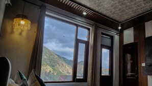 A hotel room with a bed, a large window offering a mountain view, a hanging light fixture, and a television on the wall. - Hotel Mahadev (Dharamshala)