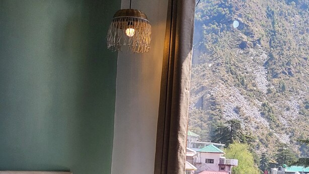 A hotel room with a bed, pillows, a hanging light fixture, and a view of a mountainous landscape with buildings. - Hotel Mahadev (Dharamshala)