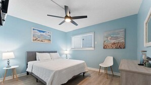 A bedroom with a bed, a ceiling fan, a window with blinds, and artwork on the walls. - Tides & Tranquility: Your Perfect Beach Getaway (North Wildwood)