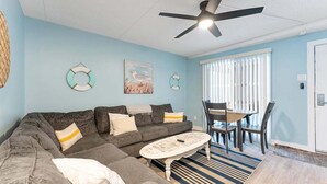 A living room with a sectional sofa, a round coffee table, a dining table with chairs, a ceiling fan, and a window with blinds. - Tides & Tranquility: Your Perfect Beach Getaway (North Wildwood)