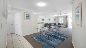 A modern living room with a glass dining table, clear chairs, and a blue rug. There is a sofa and a ceiling fan in the background.