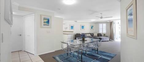 A modern living room with a glass dining table, clear chairs, and a blue rug. There is a sofa and a ceiling fan in the background.