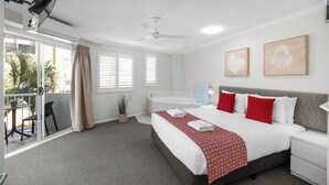 A hotel room with a bed, a bedside table, a nightstand, a ceiling fan, a window with blinds, a potted plant, and a view of the outside.