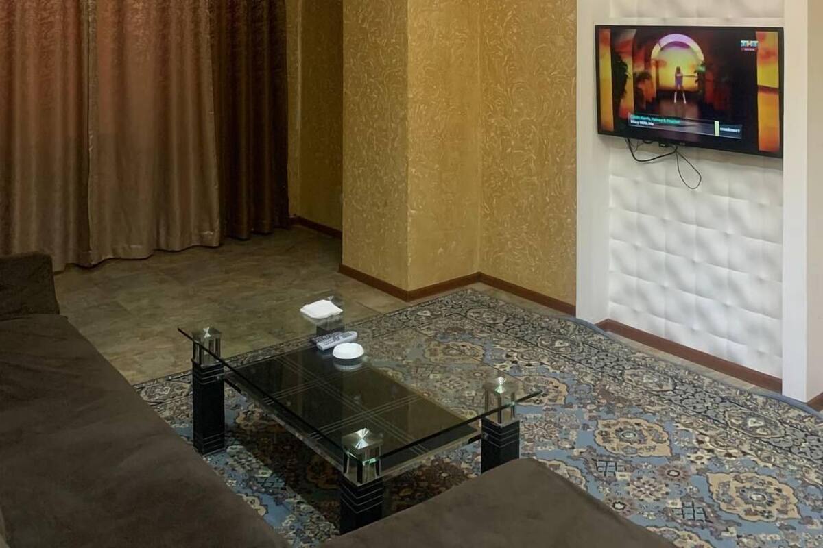 A living room with a chandelier, a television mounted on the wall, a patterned carpet, and a sofa.