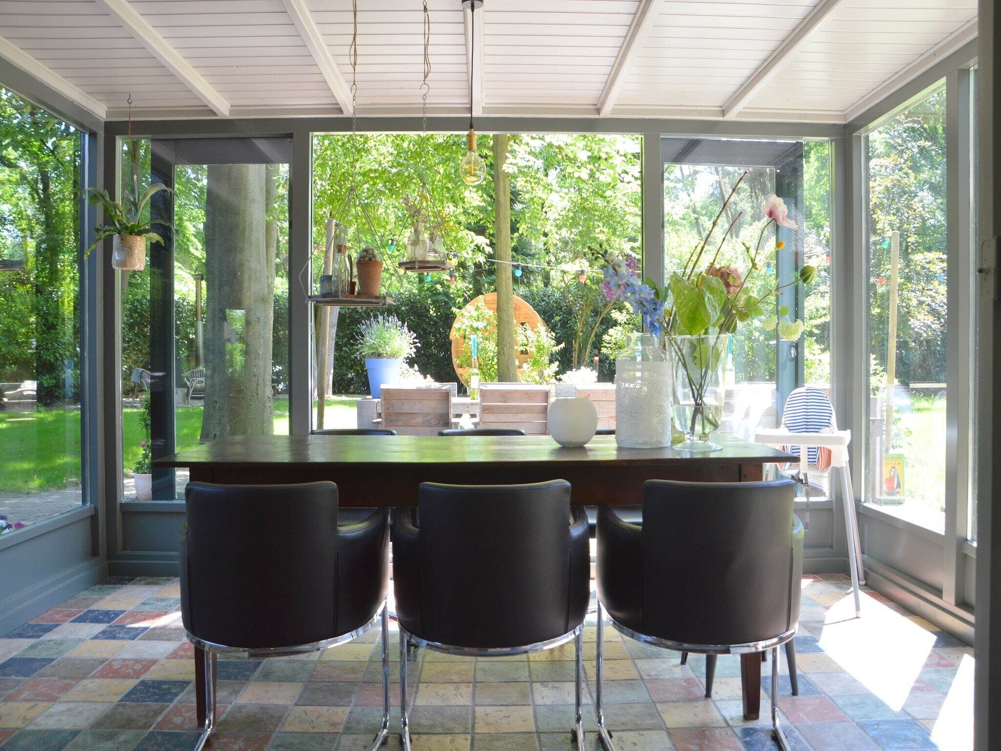 Une véranda avec une table et des chaises en verre, entourée de plantes en pot et offrant une vue sur la verdure.