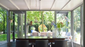 Une véranda avec une table et des chaises en verre, entourée de plantes en pot et offrant une vue sur la verdure.