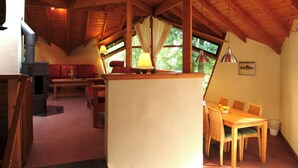 Een knus chalet met een hoog houten plafond, een open haard, een eethoek met een tafel en stoelen, en een bank.