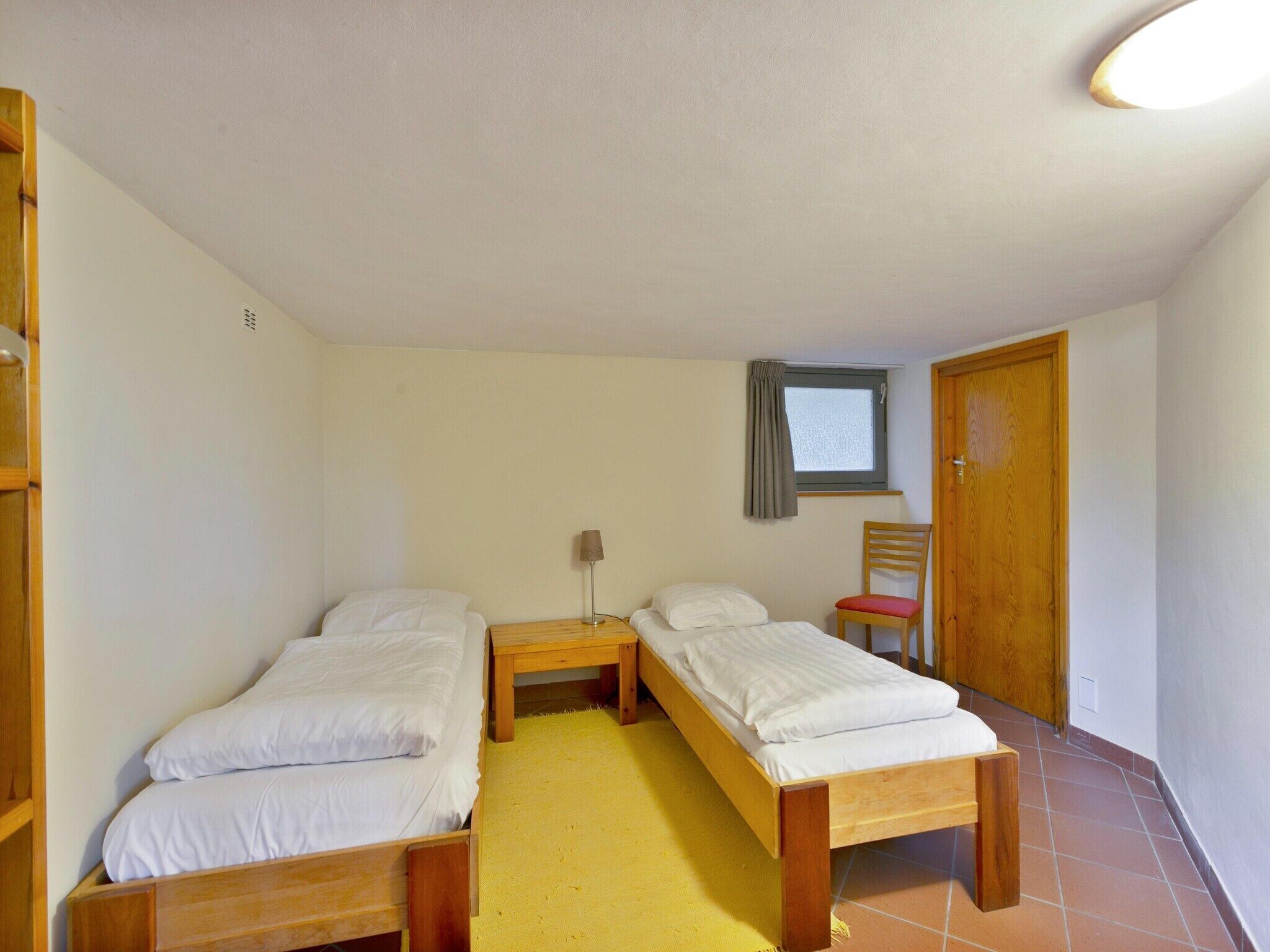 A small, clean room with two single beds, a wooden nightstand, a chair, and a window with a curtain.