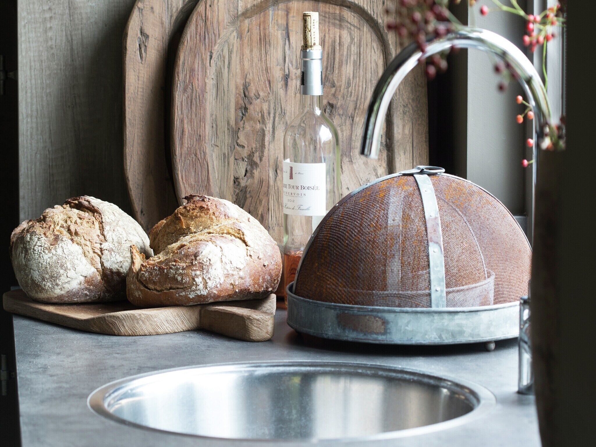 A rustic kitchen setting with a wooden cutting board, a metal sieve, and two loaves of bread.