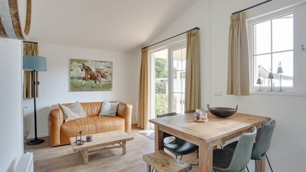 Een woonkamer met een leren bank, een houten salontafel, een eettafel met stoelen, een schilderij van een paard en een groot raam met gordijnen.